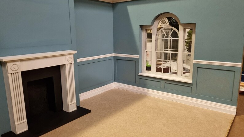 May include: A dollhouse room with blue walls, white trim, and a white fireplace. The room has a large arched window with white trim and a view of a garden.