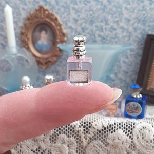 May include: A miniature clear glass perfume bottle with a square base and a silver-toned decorative cap. The bottle features a small rectangular label on the front with faint text and is sized to fit on the tip of a human finger for scale.