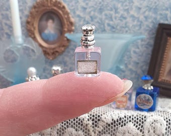 May include: A miniature clear glass perfume bottle with a square base and a silver-toned decorative cap. The bottle features a small rectangular label on the front with faint text and is sized to fit on the tip of a human finger for scale.