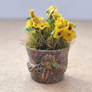 Pode incluir: Um pequeno vaso de flores cheio de flores amarelas e grama verde. O vaso &eacute; uma mistura de cores marrom, verde e dourado com uma superf&iacute;cie texturizada. As flores t&ecirc;m centros vermelho escuro.