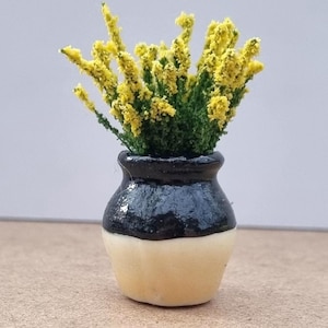 Pode incluir: Um pequeno vaso de cer&acirc;mica, preto no topo e creme na base, com flores amarelas e folhas verdes. O vaso tem cerca de 4 cm de altura, ideal para casas de bonecas ou arranjos em miniatura.