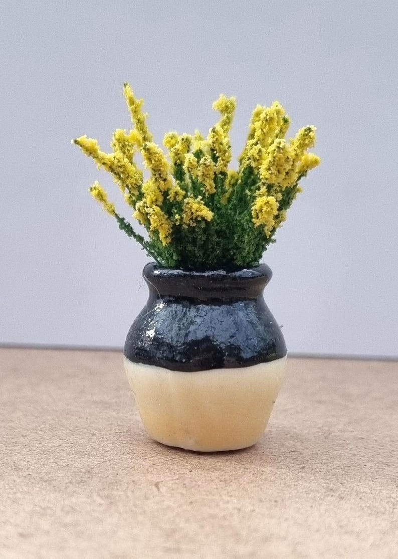 Pode incluir: Um pequeno vaso de cer&acirc;mica, preto no topo e creme na base, com flores amarelas e folhas verdes. O vaso tem cerca de 4 cm de altura, ideal para casas de bonecas ou arranjos em miniatura.