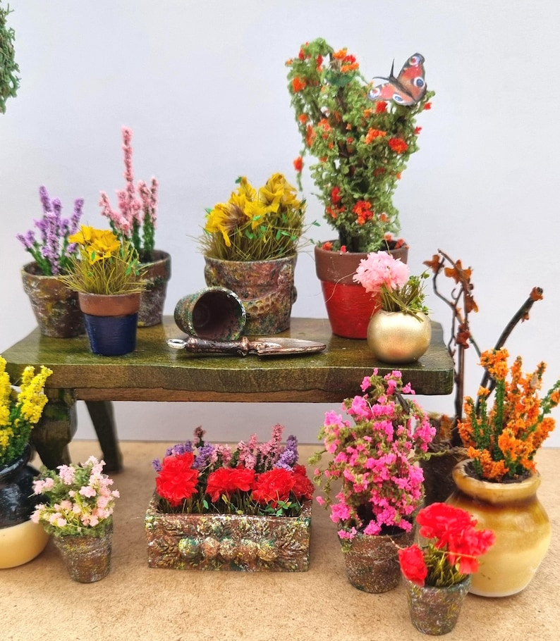 Pode incluir: Cena de jardim em miniatura com v&aacute;rias plantas e flores em vasos. Inclui um pequeno banco de madeira com uma esp&aacute;tula e um vaso virado. A cena apresenta uma variedade de flores coloridas, incluindo flores amarelas, rosas, vermelhas e laranjas, em vasos decorativos.
