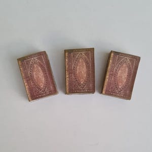 May include: Three small, antique-looking books with brown covers and gold accents. The covers have a decorative pattern with a central oval shape.