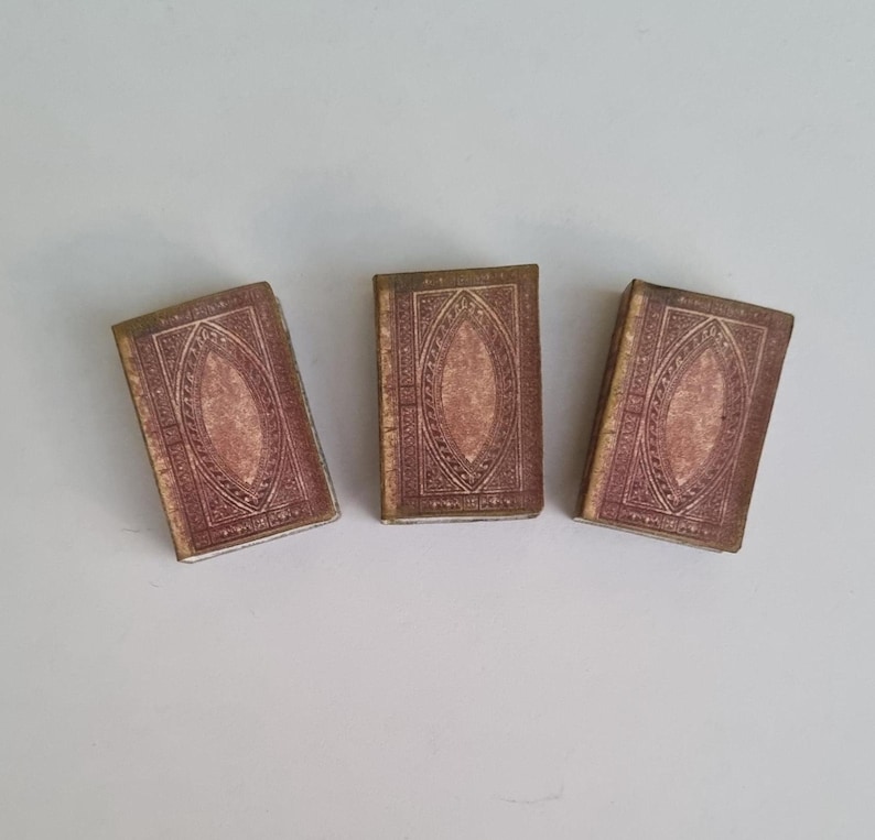 May include: Three small, antique-looking books with brown covers and gold accents. The covers have a decorative pattern with a central oval shape.