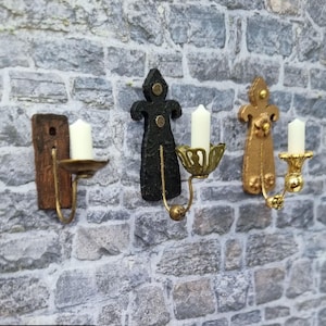 Op de afbeelding: Drie miniatuur wandkandelaars in verschillende stijlen. Eén is zwart met een eenvoudig ontwerp, één is goud met een meer sierlijk ontwerp en één is een houten plank met een eenvoudige metalen kaarshouder.