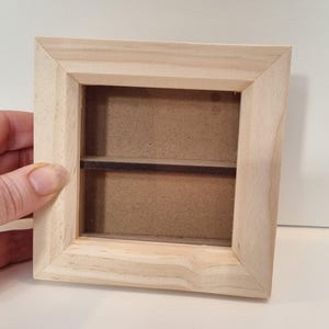 Puede incluir: Un marco de fotos de madera con un respaldo de cartón marrón y un solo estante en el interior. El marco está hecho de madera natural y tiene un diseño simple.
