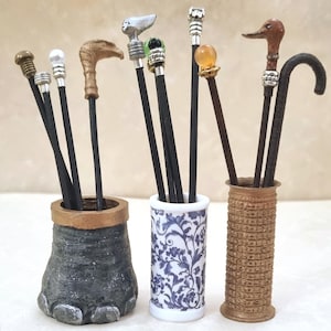 May include: A collection of miniature walking sticks with diverse decorative handles, including animal shapes and spheres, displayed in three distinct holders: a dark textured base, a white and blue floral cylinder, and a tall brown spool.