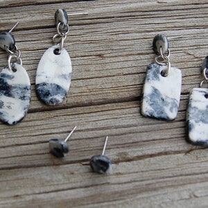 May include: A collection of handmade earrings with a marbled design. The earrings feature white and gray patterns, with silver-colored metal findings. The collection includes oval, rectangular, and square shapes, as well as a pair of stud earrings.