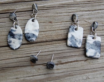 May include: A collection of handmade earrings with a marbled design. The earrings feature white and gray patterns, with silver-colored metal findings. The collection includes oval, rectangular, and square shapes, as well as a pair of stud earrings.
