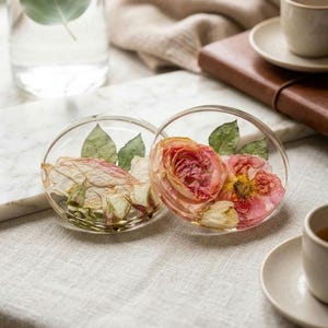 May include: Two circular clear resin coasters featuring embedded dried flowers. One coaster contains pale pink rose petals and green leaves, while the other displays vibrant pink and yellow rose blossoms with green leaves, creating a botanical display.