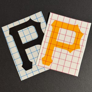 May include: Two vinyl decals featuring the letter "P". One is black, the other is orange. Both are on a white background with a grid pattern. The decals are likely for application on a car, laptop, or other surface.