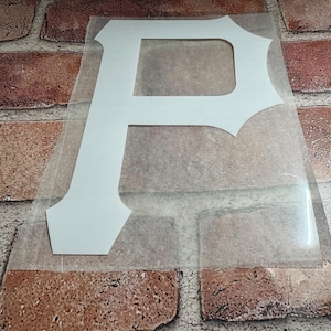 May include: A white vinyl decal in the shape of a capital letter P with a distinct serif font style. The letter is attached to a clear, rectangular adhesive transfer sheet, which is positioned flat to allow for application onto a smooth surface.