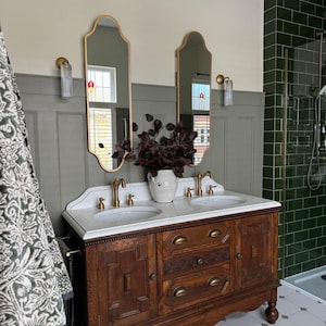 May include: A vintage wooden bathroom vanity with a marble countertop and two sinks. The vanity has brass fixtures, ornate mirrors, and a white vase with dark red foliage. The walls are painted in shades of gray and green, with a patterned tile floor.