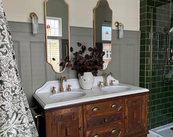 Puede incluir: Un tocador de baño de madera vintage con encimera de mármol y dos lavabos. El tocador tiene grifos de latón, espejos ornamentados y un jarrón blanco con follaje rojo oscuro. Las paredes están pintadas en tonos grises y verdes, con un suelo de baldosas estampadas.