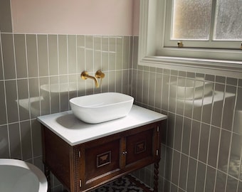 Puede incluir: Un lavabo de porcelana blanca con un grifo dorado se encuentra sobre un tocador de madera oscura con dos puertas. El tocador está sobre un suelo de baldosas de patrón blanco y negro. Las paredes están revestidas de azulejos grises y rosas.