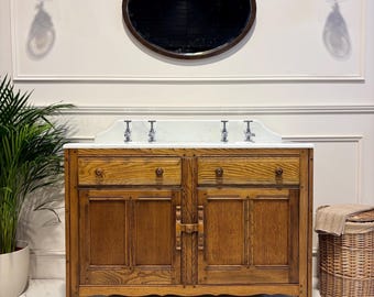 Puede incluir: Un mueble de baño de madera de estilo vintage con encimera de mármol y tres grifos. El mueble tiene dos cajones y dos puertas. Un espejo ovalado cuelga encima, con dos apliques a cada lado. También se ven una planta en maceta y una cesta de mimbre.