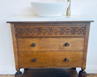 May include: A wooden bathroom vanity with a white porcelain sink and a gold faucet. The vanity has two drawers and a lower shelf. The vanity is decorated with intricate carvings.