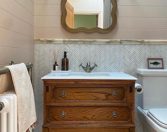 May include: A bathroom scene featuring a wooden vanity with two drawers and a white countertop. Above the vanity is a wavy-edged mirror and two sconces. The walls are painted in a light pink color with horizontal paneling. A white toilet is visible to the right.
