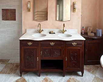 Puede incluir: Un mueble de tocador doble de madera oscura con diseños florales tallados, dos lavabos blancos y grifos dorados. Dos espejos a juego cuelgan sobre el tocador. El baño cuenta con una encimera de mármol y una pared color melocotón.