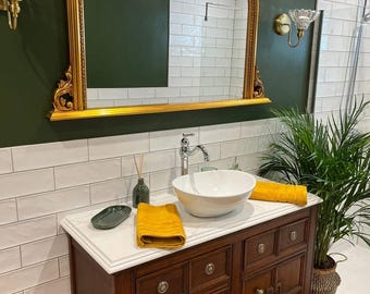 May include: A bathroom vanity with a gold-framed mirror, a white countertop, and a dark wood cabinet. The vanity features a white vessel sink, yellow towels, and decorative sconces. A potted plant adds a touch of greenery.