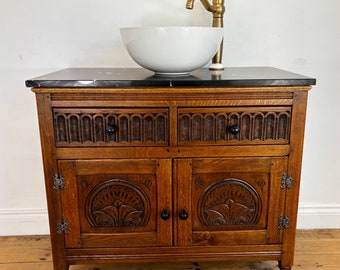 May include: A dark wood vanity with a black marble countertop and a white porcelain vessel sink. The vanity has two drawers and two doors with carved details. A gold faucet is mounted on the countertop.