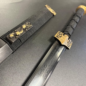 May include: A detailed image of a sword with a black handle wrapped in cord. The blade has a patterned design. The scabbard is black with gold accents and a decorative gold emblem.