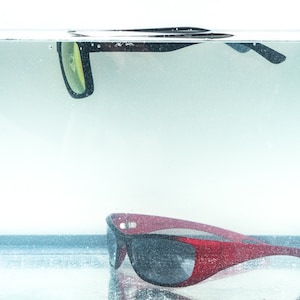 Pu&ograve; includere: Due paia di occhiali da sole, uno nero e uno rosso, parzialmente immersi nell'acqua. Gli occhiali da sole neri galleggiano sulla superficie dell'acqua, mentre gli occhiali da sole rossi riposano sul fondo del contenitore.