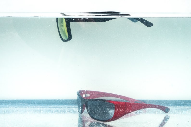 Pu&ograve; includere: Due paia di occhiali da sole, uno nero e uno rosso, parzialmente immersi nell'acqua. Gli occhiali da sole neri galleggiano sulla superficie dell'acqua, mentre gli occhiali da sole rossi riposano sul fondo del contenitore.
