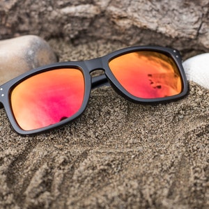 Pu&ograve; includere: Occhiali da sole neri con lenti a specchio rosse che riposano su una superficie di spiaggia sabbiosa.