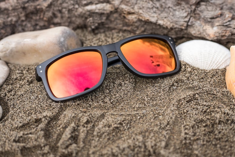 Pu&ograve; includere: Occhiali da sole neri con lenti a specchio rosse che riposano su una superficie di spiaggia sabbiosa.