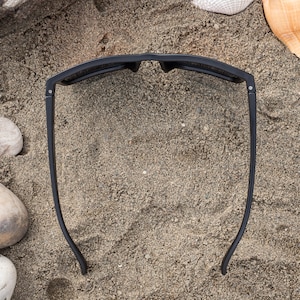 Pu&ograve; includere: Occhiali da sole neri su uno sfondo di spiaggia sabbiosa. Gli occhiali da sole sono appoggiati sulla sabbia con le lenti rivolte verso l'alto.