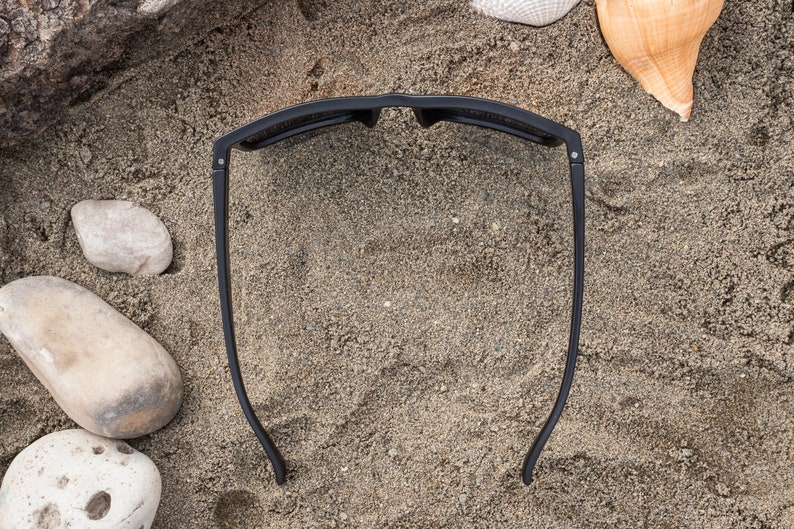 Pu&ograve; includere: Occhiali da sole neri su uno sfondo di spiaggia sabbiosa. Gli occhiali da sole sono appoggiati sulla sabbia con le lenti rivolte verso l'alto.