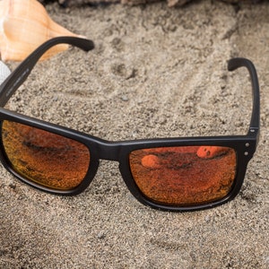 Pu&ograve; includere: Occhiali da sole neri con lenti a specchio arancioni su uno sfondo di spiaggia sabbiosa.