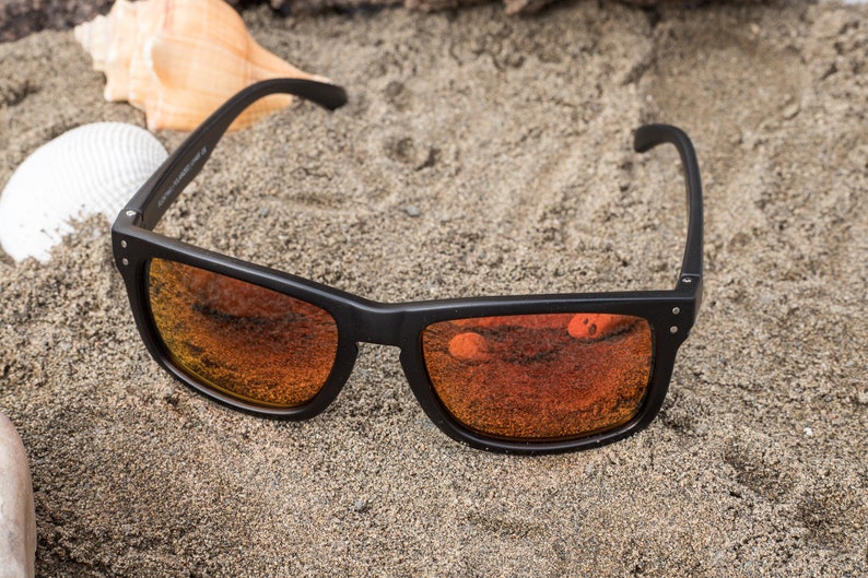 Pu&ograve; includere: Occhiali da sole neri con lenti a specchio arancioni su uno sfondo di spiaggia sabbiosa.