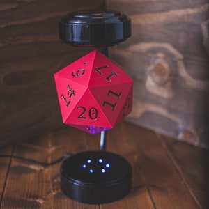 May include: A red 20-sided die levitates above a black base with blue LED lights. The die is floating in mid-air, suspended by a magnetic field.