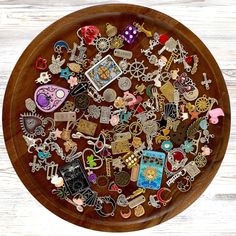 K&ouml;nnte beinhalten: Ein Holztablett gef&uuml;llt mit einer Vielzahl von Charms und Anh&auml;ngern. Die Charms sind aus Metall und haben verschiedene Designs, darunter Herzen, Sterne, Schmetterlinge und Blumen. Es gibt auch mehrere Charms mit W&ouml;rtern darauf, wie "Blessed" und "The Sun".