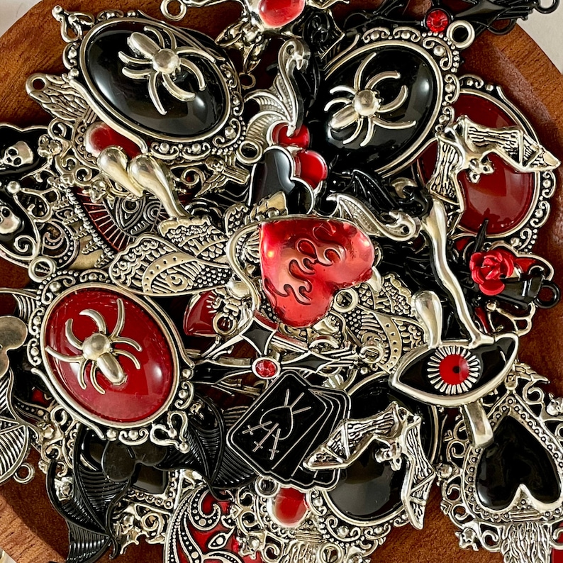 K&ouml;nnte beinhalten: Eine Sammlung von silbernen Anh&auml;nger-Anh&auml;ngern mit schwarzem und rotem Emaille. Die Anh&auml;nger zeigen Spinnen, Herzen, Augen und andere gotische Designs.