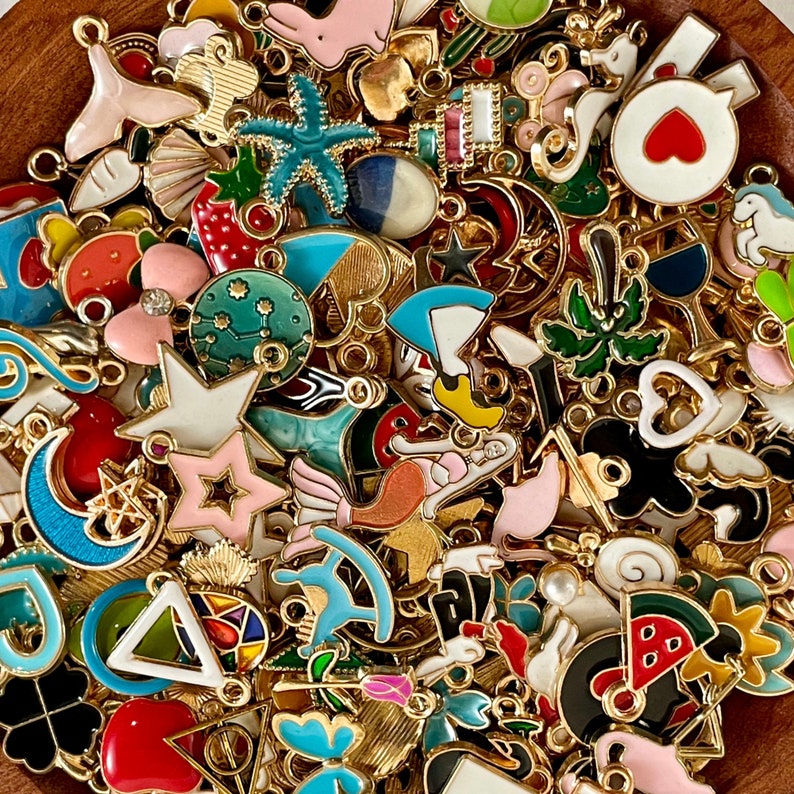 K&ouml;nnte beinhalten: Eine Sammlung bunter Emaille-Anh&auml;nger mit verschiedenen Formen und Designs, darunter Sterne, Herzen, Monde, Tiere und geometrische Muster. Die Anh&auml;nger sind aus Metall und haben eine goldene Oberfl&auml;che.