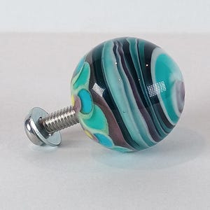 May include: A decorative glass knob with a swirling pattern of teal, purple, and white. The knob is attached to a silver metal screw and washer, suitable for furniture or cabinet hardware.