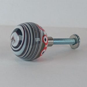 May include: A decorative cabinet knob with a round, glass head. The head features a swirl of white, black, and grey stripes, with red circular accents. It is attached to a metal screw and washer.