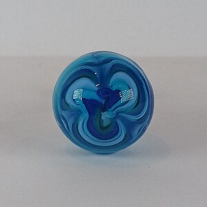May include: A spherical glass marble featuring a swirling design in shades of blue, teal, and white. The marble has a glossy surface that reflects light. The pattern evokes the imagery of ocean waves or a distant galaxy.