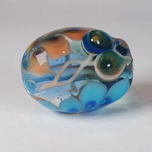 May include: A handcrafted glass bead with a swirling design. The bead features a mix of colors, including blue, orange, and green, with white accents. The bead has a smooth, rounded shape and is likely used for jewelry making or other crafts.