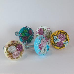 May include: A collection of five decorative cabinet knobs. Each knob features a floral design with various colors, including teal, yellow, pink, and white. The knobs have a round shape and are made of glass or ceramic.