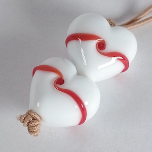 May include: A white heart-shaped pendant with a red swirl design. The pendant is made of glass and is attached to a beige cord. The pendant is a unique piece of jewelry.