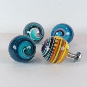May include: Four decorative glass cabinet knobs. Three knobs have swirling patterns in shades of blue and teal. The fourth knob has a striped design in yellow, orange, purple, and green, with a silver screw attached.