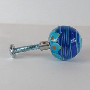 May include: A decorative cabinet knob with a round, blue glass head featuring vertical stripes and floral accents in shades of blue, turquoise, and orange. The knob is attached to a silver metal screw.