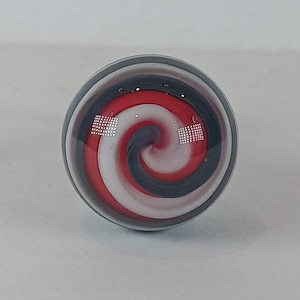 May include: A clear glass marble with a spiral design. The spiral features red, white, and dark grey bands. The marble has a dark grey band around the edge.