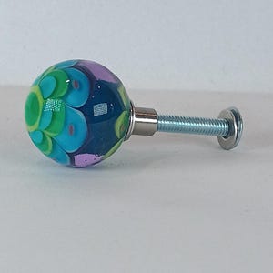 May include: A decorative glass cabinet knob with a colorful floral design. The knob is a sphere with a base of dark blue, teal, green, and purple. The knob is attached to a silver screw and washer.