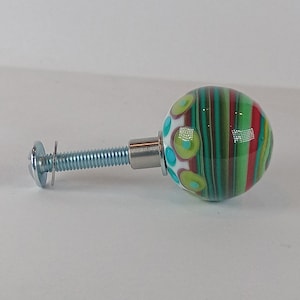 May include: A decorative glass cabinet knob with a striped pattern in green, red, and teal. The knob is round and attached to a metal screw. The knob also features white polka dots with teal and green accents.